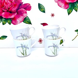 Peut inclure: Quatre tasses en céramique blanche disposées en deux piles de deux. Chaque tasse présente une illustration délicate d'une fleur d'iris violet clair avec des tiges vertes. Le fond est décoré de grandes pivoines roses et de feuilles vertes.