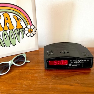 Puede incluir: Un radio despertador digital negro con una pantalla digital roja que muestra la hora 5:03. El radio despertador está sobre una superficie de madera.