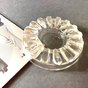 Puede incluir: Un cenicero de vidrio transparente con un borde acanalado. El cenicero tiene un centro redondo y hundido.