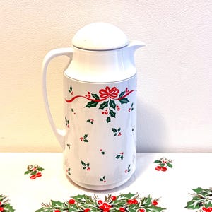 May include: White ceramic pitcher with a lid and handle, decorated with a Christmas holly and ribbon design. The pitcher has a curved handle and a spout for pouring. The holly design includes red berries and green leaves.