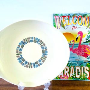 May include: A large, white ceramic serving bowl with a blue and green geometric design around the center. The bowl is on a wooden surface and a sign that says "Welcome to Paradise" with pink flamingos is in the background.