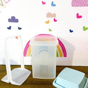 May include: A clear plastic container with a pink lid, a light blue container, and a white plastic utensil holder are on a wooden surface. The background features a rainbow and colorful heart and cloud stickers.