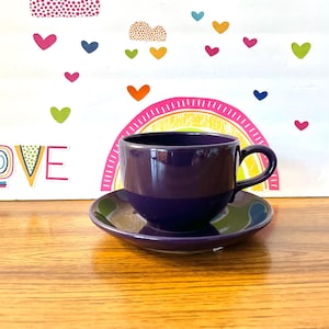 Puede incluir: Un juego de taza y platillo de color morado. La taza tiene una forma redondeada y un asa. El platillo es de forma redonda simple.