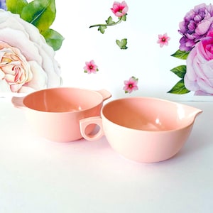 Puede incluir: Dos cuencos de color rosa claro. Un cuenco tiene un asa y un pequeño pico. El otro cuenco tiene un asa. Los cuencos están sobre una superficie blanca con un fondo floral con flores rosas y moradas y hojas verdes.