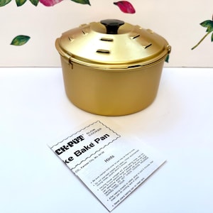 May include: A gold-colored metal slow cooker bake pan with a matching lid featuring a black knob and ventilation slots. The pan is accompanied by a white instructional pamphlet titled CK-POT SLOW COOKER Bake Bake Pan with printed text and hints.