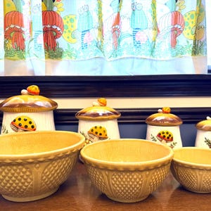 Puede incluir: Una colección de utensilios de cocina de cerámica vintage, que incluye cuencos y botes. Los cuencos son de color amarillo claro con un patrón texturizado. Los botes tienen tapas en forma de hongo y diseños de hongos y mariquitas.