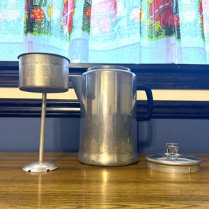以下が含まれることがあります： ヴィンテージのアルミニウム製コーヒーパーコレーターセット。セットには、黒いハンドルが付いた背の高い円筒形のポット、スタンド付きの穴あきフィルターバスケット、透明なガラスノブ付きの蓋が含まれます。アイテムは木製の表面に表示されます。