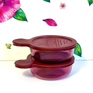 Puede incluir: Dos cuencos apilados, translúcidos, de color burdeos, con tapas a juego. Cada cuenco tiene una extensión en forma de asa. Las tapas tienen texto. El fondo es blanco con diseños florales y de hojas.