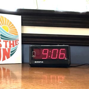 Puede incluir: Un reloj despertador digital negro que muestra las 9:06 en números rojos está sobre una superficie de madera. Junto a él, una impresión en lienzo cuadrada con las palabras "HERE COMES THE SUN" en naranja y azul, con un diseño de sol y olas.