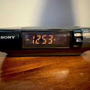 May include: A black Sony digital alarm clock showing the time 12:53 AM in orange digits. The clock has buttons for alarm, mode, and radio functions. The clock is on a wooden surface.