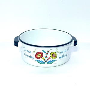 May include: White enamelware bowl with black handles and rim. The bowl features a floral design in red, yellow, and blue, with text in a script font. A vintage kitchenware item.