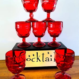 May include: A set of seven red glass goblets with a faceted, geometric texture on the bowls and stems. The glasses are arranged in a pyramid formation on a black rectangular base that features the word "cocktail" printed in black lettering.