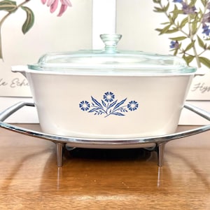 May include: A white rectangular casserole dish with a blue floral pattern on the front, featuring a clear glass lid and a chrome metal serving cradle with four legs. The dish has integrated side handles and a smooth, glossy finish throughout.