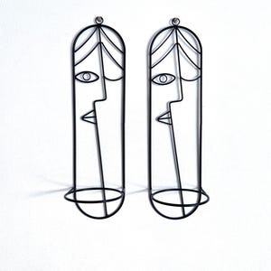 May include: Two black metal wall art pieces, each featuring a minimalist face design. The art pieces have a long, oval shape with a circular base and a small loop at the top for hanging. The faces have simple line-drawn features.