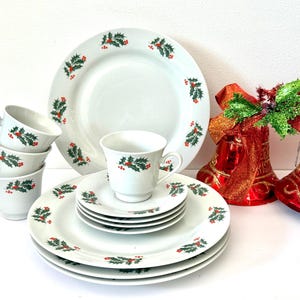 May include: A white Christmas dinnerware set with a holly berry design. The set includes plates, saucers, and cups. Two red Christmas bells with gold trim and bows are in the background.