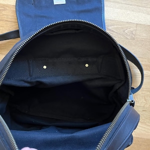 May include: Open view of a black backpack with a top flap and zipper closure. The interior is black fabric. The backpack has two shoulder straps and a top handle. The zipper is silver-toned.