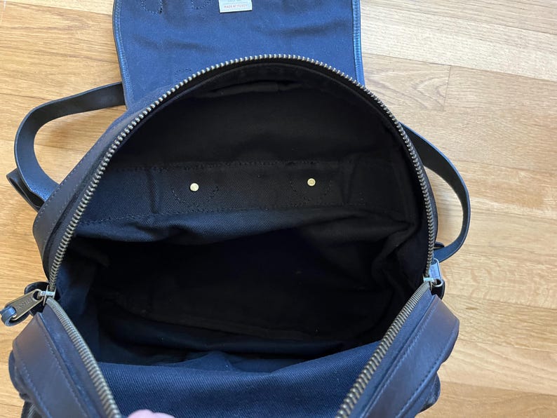 May include: Open view of a black backpack with a top flap and zipper closure. The interior is black fabric. The backpack has two shoulder straps and a top handle. The zipper is silver-toned.