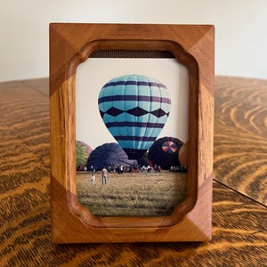 May include: A wooden picture frame with a photograph of hot air balloons. The frame is made of light brown wood and has a decorative, curved edge. The photograph features a large blue and black hot air balloon, with other balloons in the background.