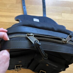 May include: Black leather saddlebag with a zippered main compartment and multiple smaller pockets. The bag features silver-toned zippers and buckles, and adjustable straps. The bag is designed to be mounted on a bicycle or motorcycle.
