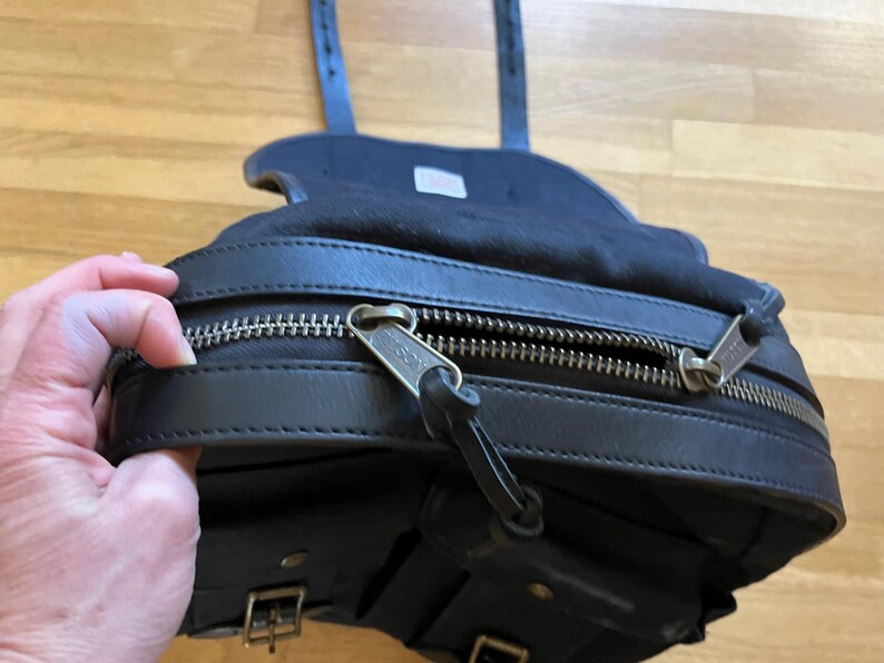 May include: Black leather saddlebag with a zippered main compartment and multiple smaller pockets. The bag features silver-toned zippers and buckles, and adjustable straps. The bag is designed to be mounted on a bicycle or motorcycle.