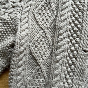 May include: Close-up of a light gray knitted sweater with intricate cable and diamond patterns. The textured knitwear displays a variety of stitches, creating a cozy and detailed design. The sweater's neutral color makes it versatile.