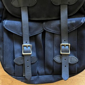 May include: Black canvas backpack with two front pockets, secured by leather straps and brass buckles. The backpack features a top flap and adjustable shoulder straps. The design is classic and durable, suitable for carrying various items.
