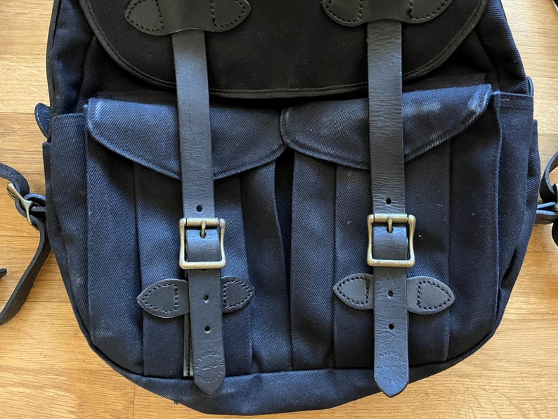 May include: Black canvas backpack with two front pockets, secured by leather straps and brass buckles. The backpack features a top flap and adjustable shoulder straps. The design is classic and durable, suitable for carrying various items.