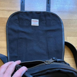 May include: Black canvas messenger bag with a leather flap and straps. The bag has a zippered main compartment and a small tag that reads "FILSON MADE IN USA". The bag is open, revealing the interior.