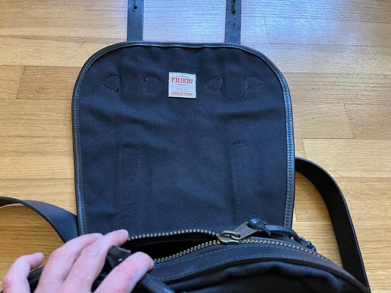 May include: Black canvas messenger bag with a leather flap and straps. The bag has a zippered main compartment and a small tag that reads "FILSON MADE IN USA". The bag is open, revealing the interior.