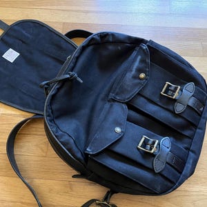 May include: A black backpack with a flap and two front pockets secured by leather straps and metal buckles. The backpack has a zippered main compartment and adjustable shoulder straps. A black flap with a white tag is open next to the backpack.