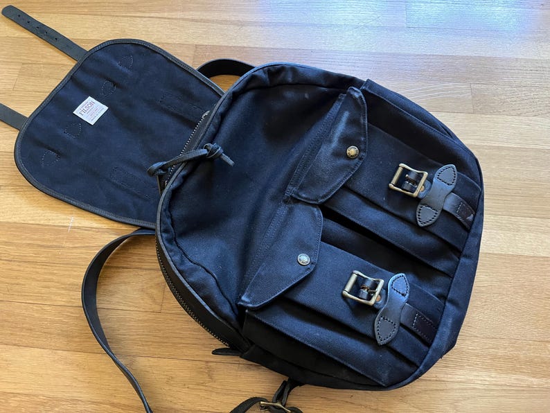May include: A black backpack with a flap and two front pockets secured by leather straps and metal buckles. The backpack has a zippered main compartment and adjustable shoulder straps. A black flap with a white tag is open next to the backpack.
