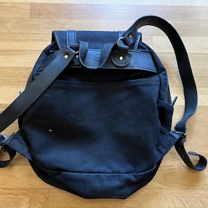 May include: Black canvas backpack with leather straps and accents. The backpack features a flap closure, adjustable shoulder straps, and a front pocket. The bag has a classic, outdoorsy design, suitable for everyday use.