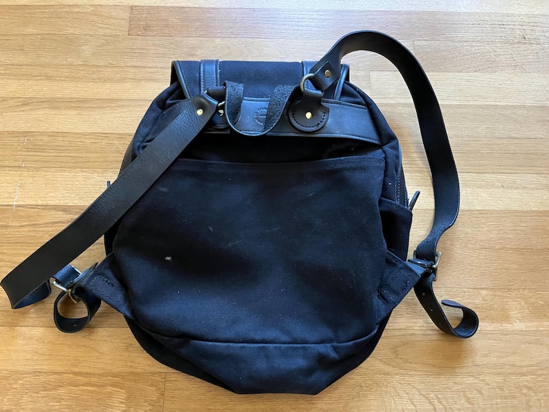 May include: Black canvas backpack with leather straps and accents. The backpack features a flap closure, adjustable shoulder straps, and a front pocket. The bag has a classic, outdoorsy design, suitable for everyday use.