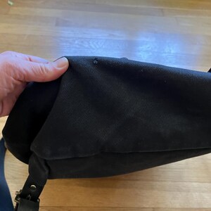 May include: Black canvas messenger bag with a flap closure, leather straps, and metal hardware. The bag has a shoulder strap and a handle. The bag is shown on a wooden surface.