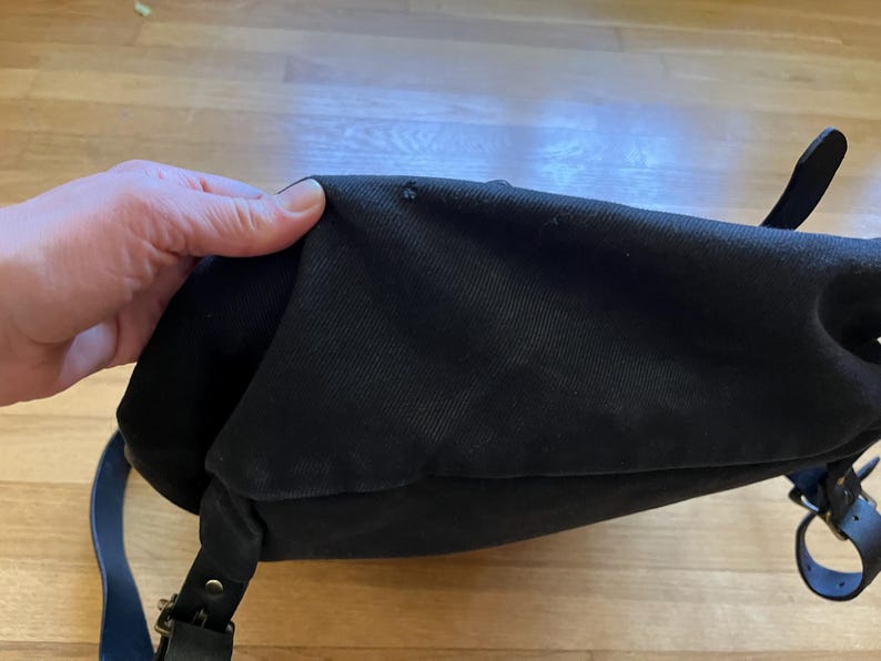 May include: Black canvas messenger bag with a flap closure, leather straps, and metal hardware. The bag has a shoulder strap and a handle. The bag is shown on a wooden surface.