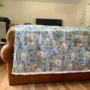 May include: A rectangular piece of fabric featuring a light blue background with a repeating floral pattern of cream-colored blossoms, dark green leaves, and small blue accents. The fabric has a raw, unfinished white edge along the bottom.