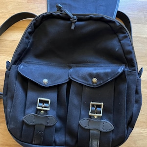 May include: Black canvas backpack with two front pockets secured by leather straps and metal buckles. The backpack has adjustable shoulder straps and a top handle. The bag features a zippered main compartment and a flap closure.