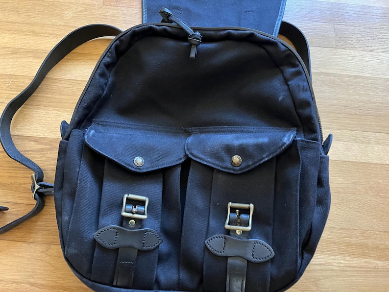 May include: Black canvas backpack with two front pockets secured by leather straps and metal buckles. The backpack has adjustable shoulder straps and a top handle. The bag features a zippered main compartment and a flap closure.