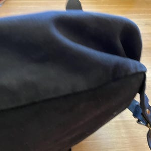May include: Black satchel bag with a curved top and a leather strap with a brass buckle. The bag appears to be made of a soft, dark fabric.