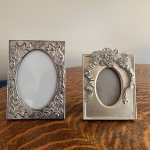 May include: Two silver-toned picture frames. The frame on the left has an oval opening and is decorated with an ornate floral and leaf pattern. The frame on the right has an oval opening and is decorated with roses and a ribbon.
