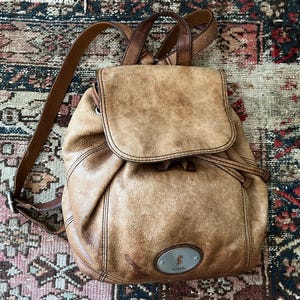 May include: A tan leather backpack with a flap closure and drawstring. The bag has adjustable straps and a small metal Fossil logo. The leather has a distressed appearance, with visible wear and tear. The backpack is sitting on a patterned rug.