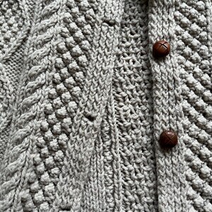 May include: Close-up of a light gray knitted cardigan with intricate cable and textured stitch patterns. The cardigan features two brown, round buttons. The overall design suggests a cozy, handmade garment.