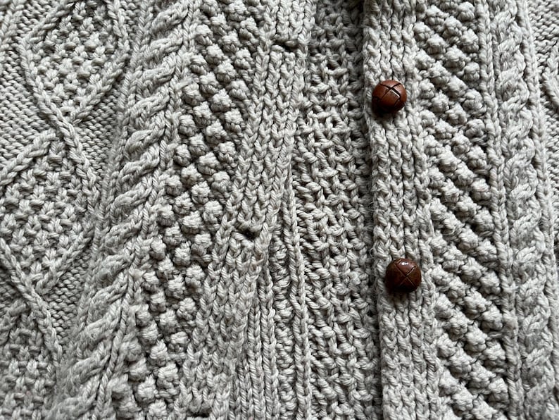 May include: Close-up of a light gray knitted cardigan with intricate cable and textured stitch patterns. The cardigan features two brown, round buttons. The overall design suggests a cozy, handmade garment.