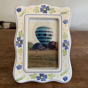 May include: A ceramic photo frame with a scalloped edge, decorated with blue floral designs and yellow accents. The frame holds a picture of hot air balloons in a field. The frame is approximately 6 inches tall and 4 inches wide.