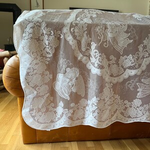 May include: A white lace tablecloth with a detailed floral and angel design. The delicate fabric drapes over a brown leather sofa, showcasing the intricate patterns and textures. The tablecloth's edges are scalloped, adding a decorative touch.