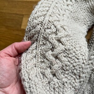 May include: Close-up of a beige cable-knit sweater sleeve. The textured knitwear features intricate cable patterns and a ribbed cuff. The soft, warm material suggests a cozy, handmade garment, perfect for cooler weather.