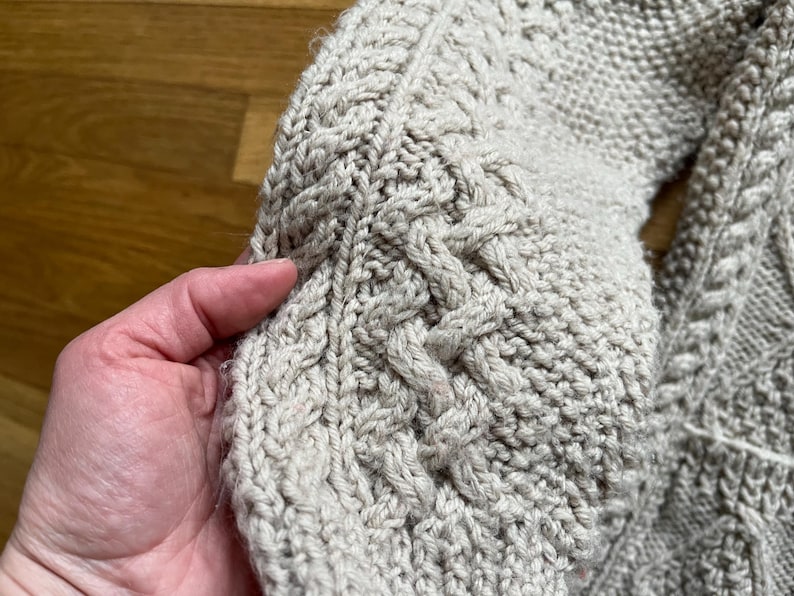 May include: Close-up of a beige cable-knit sweater sleeve. The textured knitwear features intricate cable patterns and a ribbed cuff. The soft, warm material suggests a cozy, handmade garment, perfect for cooler weather.
