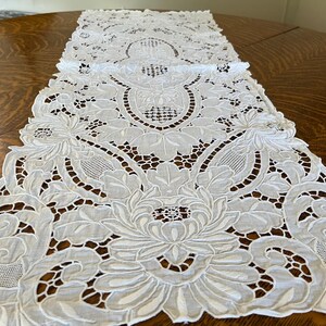 May include: A white, intricately embroidered table runner with a floral design. The runner is made of a delicate fabric and features detailed cutwork and scalloped edges. The runner is placed on a round, wooden table.
