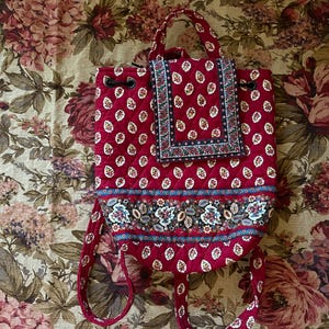 May include: Red quilted backpack with a floral pattern and a matching flap pocket. The backpack features a drawstring closure, adjustable straps, and a top handle. The design includes a blue and floral patterned band across the bottom.