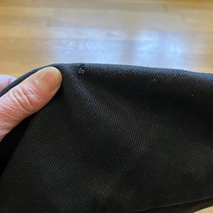 May include: Close-up of black fabric with a small area of damage. The fabric is held by a hand, showing the texture and color of the material. The background is a light brown wooden surface.